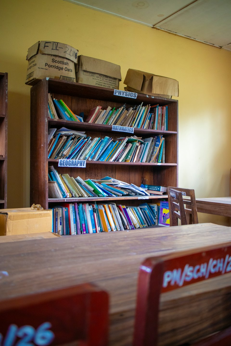 A cluttered wooden bookshelf filled with books and boxes in a classroom.